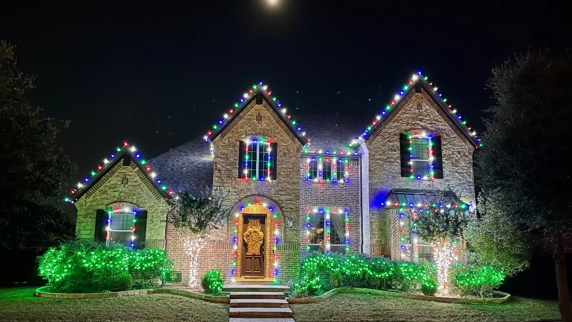 Stone two-story home with blue and white Christmas lights and wrapped trees
